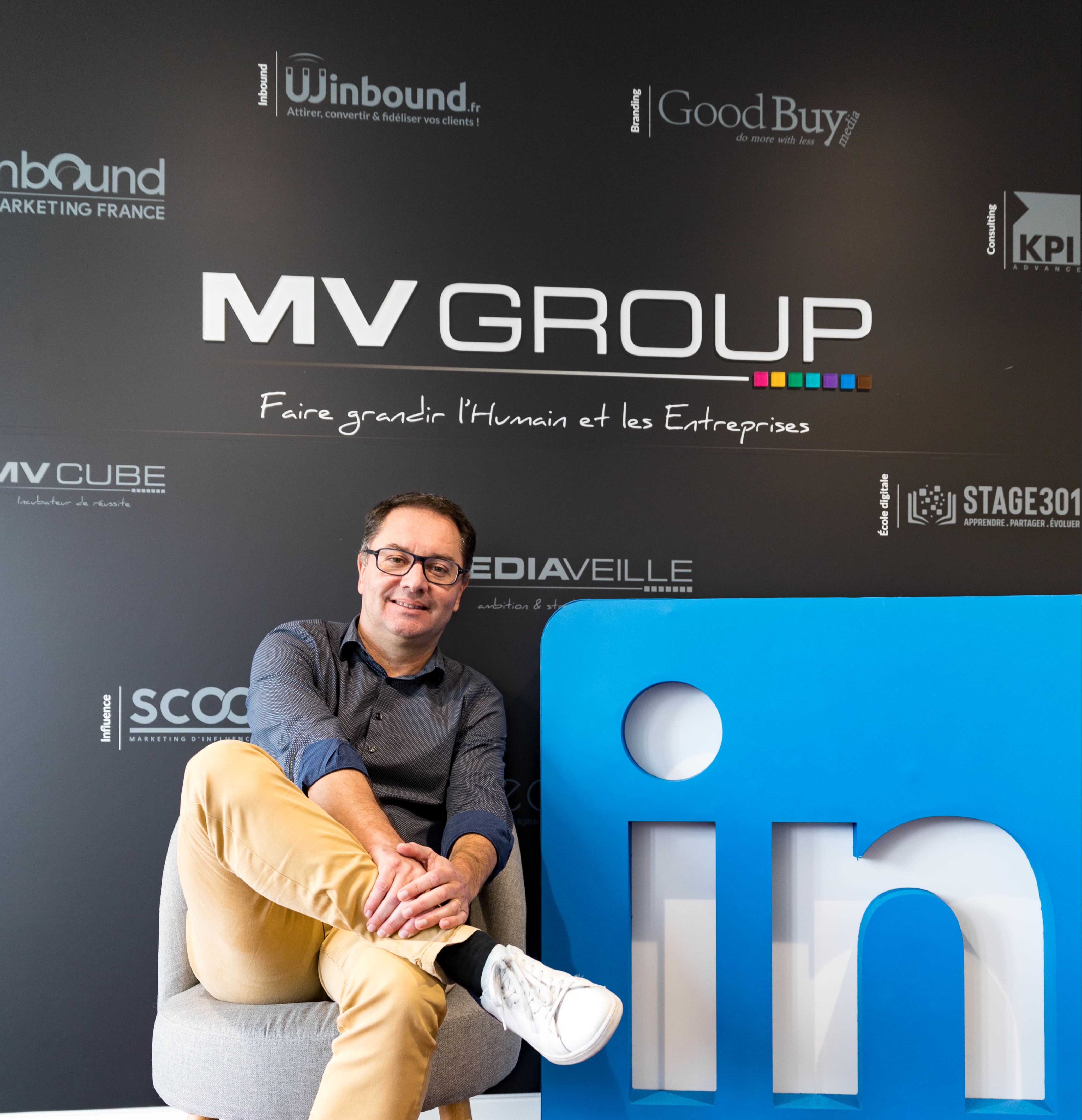 Olivier Méril assis sur une chaise devant logo MV Group et logos de filiales